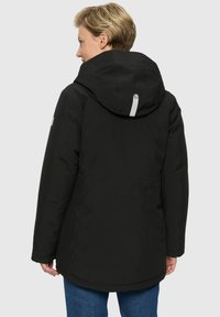 Persoon met kort blond haar, gekleed in een zwarte hoodie met een reflecterende strook op de capuchon, met de rug naar de camera tegen een effen achtergrond.