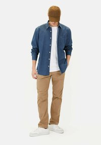 Dunkelblaue Jeanshemd, weißes T-Shirt, sandfarbene Hose und weiße Sneaker. Eine braune Kappe rundet das Outfit ab.