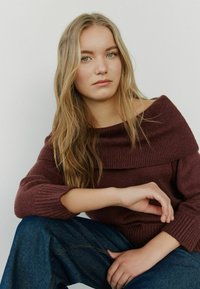 Burgundy off-the-shoulder knit sweater with ribbed texture, featuring wide sleeves and paired with dark denim trousers.