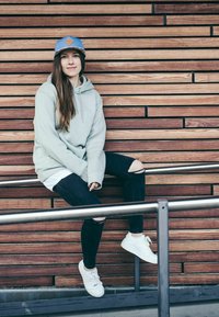 Light grey hoodie, ripped black jeans, white sneakers, and a blue cap with a logo, seated on a railing against a wooden wall.