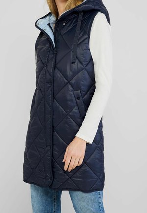 Žena v dlouhé námořnicky modré prošívané vestě s kapucí přes bílé tričko s dlouhým rukávem a modré džíny, stojící s rukou opřenou o vestu.