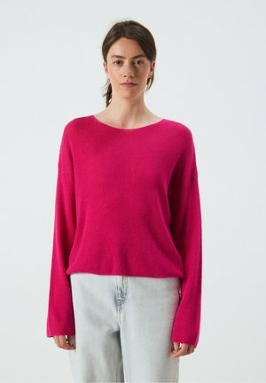Frau mit dunklen Haaren trägt einen knallpinken Langarmpullover und hellblaue Jeans, steht vor einem einfarbigen hellen Hintergrund.