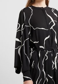 Schwarzes Kleid mit weißem abstraktem Linienmuster, langen Ärmeln, Knopfverschluss und einem Bindegürtel in der Taille. Glatter, leichter Stoff.