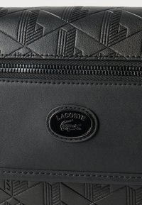 Sac noir texturé avec motif géométrique en relief, une fermeture éclair horizontale et une plaque ovale noire avec le logo Lacoste et l'emblème du crocodile.