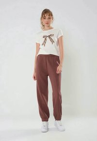 Kvinna klädd i en vit kortärmad skjorta med leopardmönstrad rosett och högmidjade bruna joggerbyxor, tillsammans med vita sneakers, som står mot en neutral vägg.