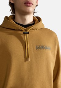 Sudadera con capucha de color mostaza, con impresión de logo, que presenta cordones redondos con puntas metálicas y una tela suave y texturizada.