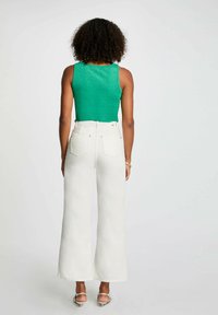 Top verde scintillante abbinato a jeans bianchi a vita alta con gamba larga. L'outfit presenta una texture liscia e un design minimalista.
