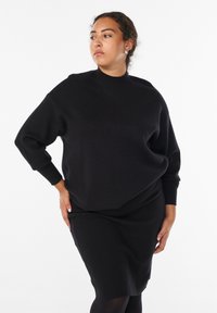 Kobieta z kręconymi włosami, ubrana w czarny sweter z długim rękawem i dopasowaną spódnicę, pozująca z rękami na biodrach, patrząca w bok.