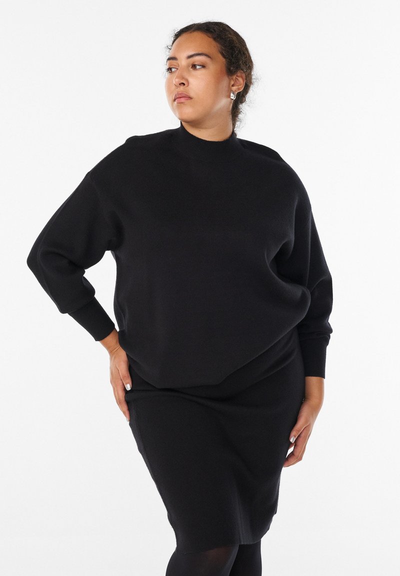 Kobieta z kręconymi włosami, ubrana w czarny sweter z długim rękawem i dopasowaną spódnicę, pozująca z rękami na biodrach, patrząca w bok.