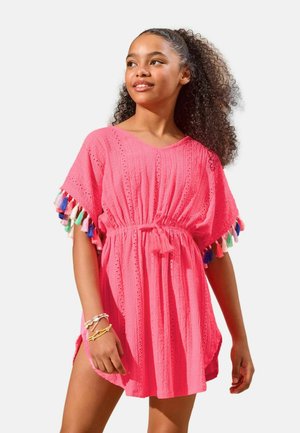 Dievča s kučeravými vlasmi oblečené v ružových šatách s farebnými strapcami na rukávoch a viacerými náramkami, jemne sa pozerá do strany a usmieva sa.