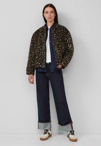 Vrouw staat met een panterprint jack, donkerblauwe wijde jeans met omslag, witte sneakers, een spijkerblouse en een wit T-shirt tegen een effen achtergrond.