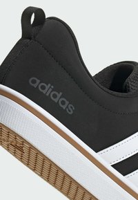 Čierny teniska s hladkým gumovým zvrškom, bielymi doplnkami a textúrovanou podrážkou. Má slučku pre ľahké obliekanie a šedé logo „adidas“.