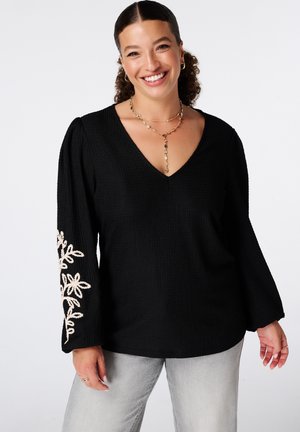 Femme souriante portant un pull noir col en V avec une broderie florale beige sur les manches et un pantalon gris clair, accessoirisée de bijoux en or.