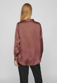 Blus med långa ärmar i mörk mauve satin, med krage, slät textur, rundad fåll och en-knapps manschetter. Svarta byxor delvis synliga.