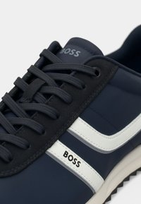 Ténis azul marinho com riscas brancas, apresentando uma parte superior texturizada, atacadores planos e a marca "BOSS" impressa no lado.