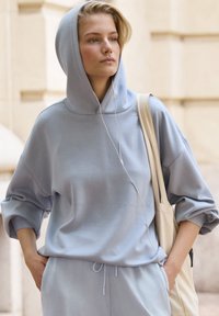 Sudadera azul claro con capucha, de corte holgado y mangas remangadas. Llevado con pantalones a juego y un bolso de mano color crema. Auriculares conectados.