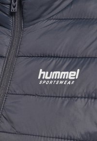 Nahaufnahme einer dunkel gepolsterten Sportjacke mit sichtbarem Reißverschluss und weißem „hummel SPORTSWEAR“-Logo auf dem Stoff.