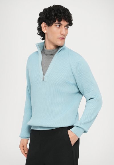 Pull bleu clair à fermeture éclair avec texture côtelée et manches longues, porté sur un col roulé gris. Associé à un pantalon noir.
