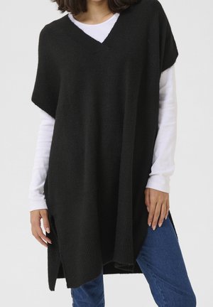 Vrouw die een zwarte gebreide mouwloze poncho draagt over een wit shirt met lange mouwen en blauwe spijkerbroek, staand tegen een effen achtergrond.
