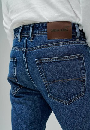 Jean en denim bleu foncé avec une finition texturée, présentant des coutures visibles et une étiquette en cuir marron marquée "SALSA JEANS" sur la ceinture.