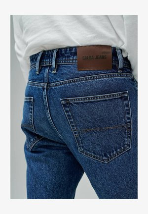 Jean en denim bleu foncé avec une finition texturée, présentant des coutures visibles et une étiquette en cuir marron marquée "SALSA JEANS" sur la ceinture.