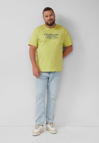 Homme barbu avec les cheveux attachés portant un t-shirt jaune "Outdoorsy", un jean bleu clair et des baskets blanches, se tenant devant un mur uni.