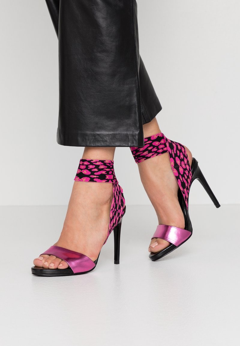 Jambes féminines portant un pantalon en cuir noir coupé et des sandales à talons hauts avec une bride avant métallisée rose et un enroulement de cheville en imprimé léopard rose et noir.