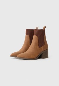 Stivali alla caviglia in suede marrone con bordi in maglia a coste e tacchi a blocco, vista angolata frontale su sfondo bianco.