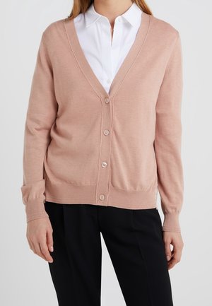 Vrouw in een lichtroze knopen cardigan over een witte blouse met kraag en zwarte broek, staand tegen een effen achtergrond.