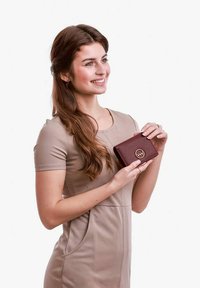 Burgundy leather wallet with a smooth texture, featuring a gold circular logo. Model in fitted beige dress holds the wallet with both hands.