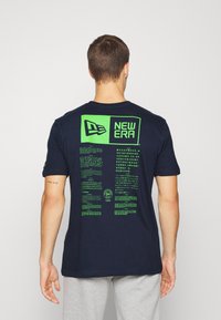 Námořnická modrá bavlněná tričko s velkým zeleným grafickým motivem na zadní straně, obsahujícím text a logo, krátké rukávy a klasický design kulatého výstřihu.