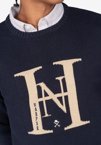 Jersey de punto azul marino con un gran logotipo beige "HN", tejido texturizado, cuello acanalado y pequeño detalle de texto "HARPER" en la parte inferior.