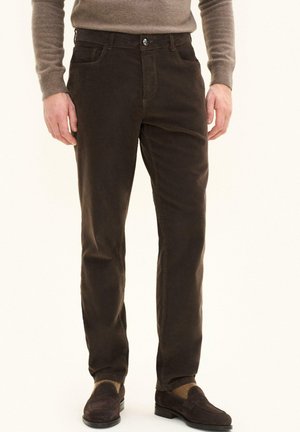 Man draagt bruine ribfluwelen broek, bruine loafers en een beige trui met lange mouwen, staand tegen een effen achtergrond.