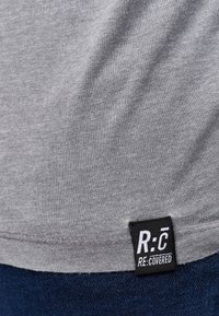 Šedé tričko vyrobené z měkké bavlny, na spodním švu s černou etiketou a bílým textem "R:C RE:COVERED", hladká struktura.