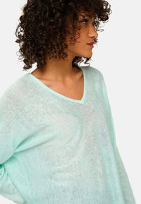 Haut transparent vert menthe clair à col en V, avec des manches longues, une coupe ample et un tissu doux texturé, mettant en valeur une silhouette décontractée.