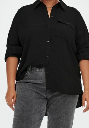 Persona con curvas que lleva una camisa negra holgada de botones con las mangas remangadas y unos vaqueros de talle alto en gris oscuro.