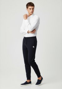Marineblaue Jogginghosen mit seitlichen Taschen, elastischem Bund und gerippten Bündchen. Kombiniert mit einem taillierten weißen Hemd und marineblauen Slipschuhen.