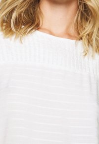 Bílý top s texturovaným designem, který má jemné vodorovné pruhy a široký výstřih. Tkanina vypadá lehce a měkce.