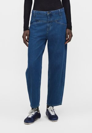 Personne portant un jean bleu taille haute, coupe ample, avec une ceinture asymétrique unique, un haut noir à manches longues et des baskets noires avec des lacets blancs.
