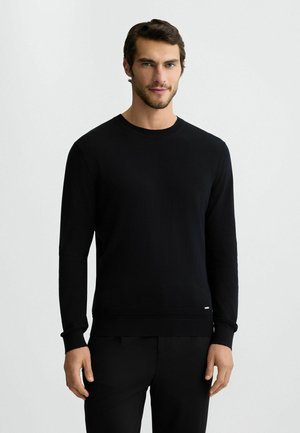 Suéter negro de manga larga y cuello redondo en una suave tela de punto. Presenta puños y dobladillo acanalados, con un pequeño logotipo en la parte inferior.