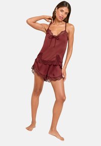 Frau steht barfuß in einem burgunderfarbenen Satin-Camisole und Short-Set mit Spitzenbesatz, während sie mit einer Hand hinter ihrem Kopf und der anderen an ihrer Seite posiert.