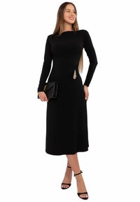 Czarna sukienka z długim rękawem, dopasowaną górą i rozszerzanym dołem. Posiada złoty detal w talii oraz gładką fakturę materiału.