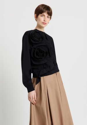 Donna con capelli castani corti che indossa una maglia nera con grandi dettagli floreali in tessuto e una gonna plissettata beige, in piedi contro uno sfondo bianco.