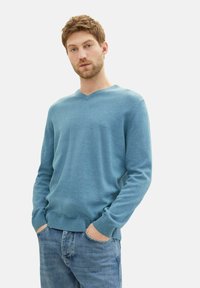 Hellsblauer V-Ausschnitt-Pullover aus weichem Material, mit langen Ärmel und gerippten Bündchen, kombiniert mit hellblauen Jeans.