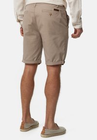 Shorts en coton beige avec des poignets roulés, des poches sur la taille et une poche au dos avec fermeture à bouton. Portés avec des chaussures légères à enfiler.