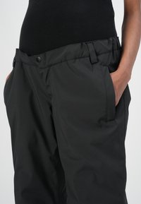 Zwarte waterdichte broek met een gladde afwerking, voorzien van zijzakken en een tailleband met riemlussen en drukknoopsluitingen.