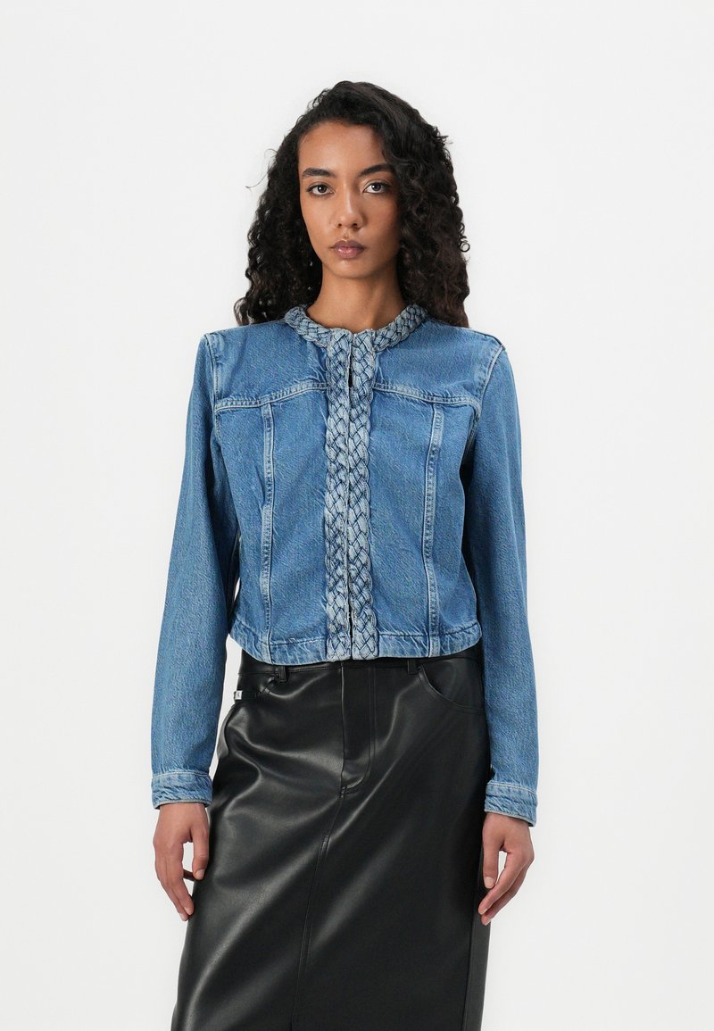 Veste en denim avec un devant tressé, présentant des manches longues, une coupe courte et un tissu bleu. Associée à une jupe en cuir noir élégante.