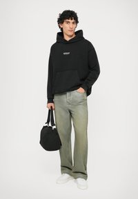 Hoodie noir avec poche avant, jean large déchiré vert clair, sac noir et baskets blanches. Cheveux bouclés, fond neutre.