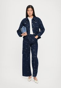 Tumed sinine denim jakk ja püksid, millel on tähe muster. All on valge T-särk, käes on väike sinine kott ja jalas kreemivärvi kingad.