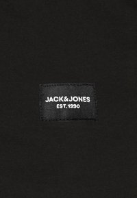 Černá bavlněná látka s obdélníkovou tkanou etiketou s bílým textem: "JACK & JONES EST. 1990." Žádné další vzory ani textury nejsou viditelné.